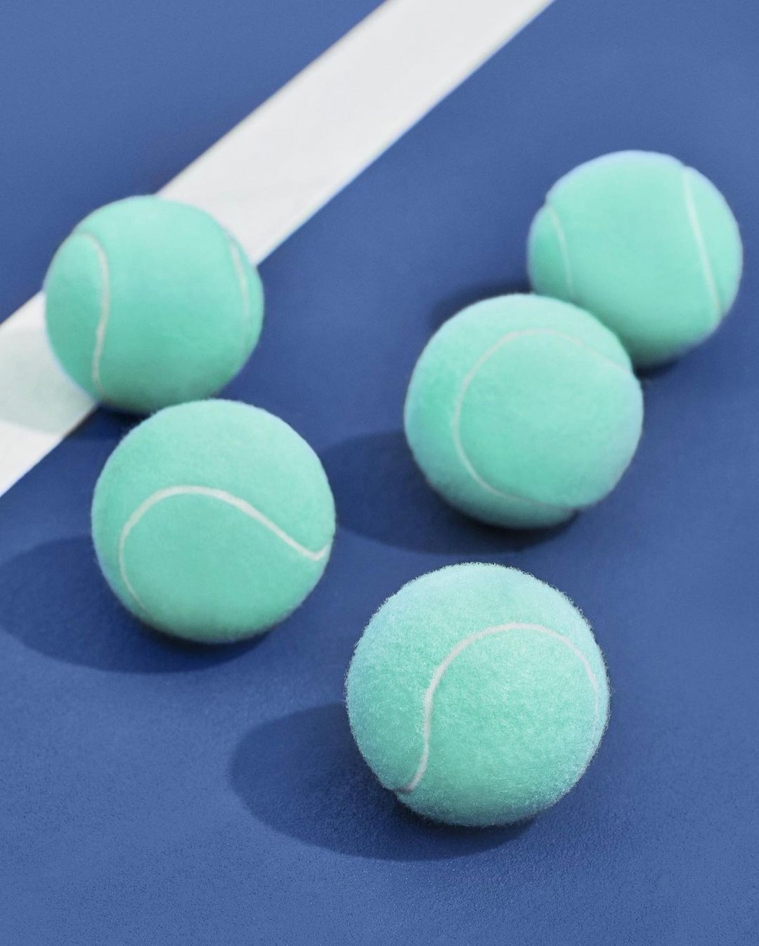 Five light blue-green tennis balls arranged on a blue surface with a white line. Sports equipment on a solid background with shadow detail.