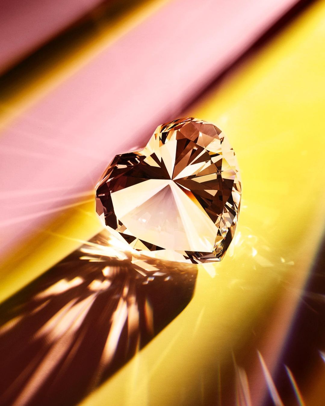 Heart-shaped diamond on a reflective surface with yellow and pink lighting creating refracted shadows and highlights, studio shot.