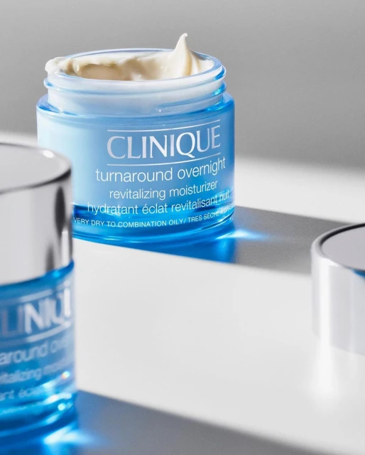 Close-up product shot of Clinique Turnaround Overnight revitalizing moisturizer in a blue glass jar with white cream on a white surface.