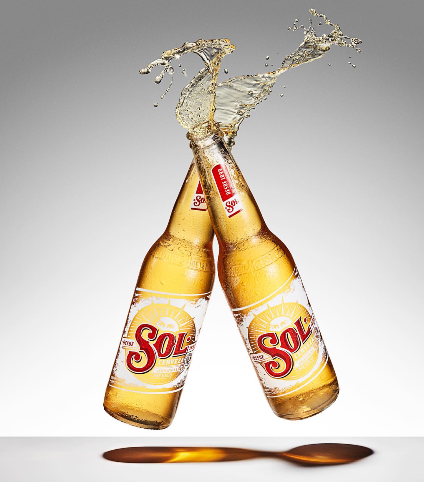 Two Sol beer bottles clink together in a celebratory toast, creating a splash against a gradient grey background and a wooden surface.