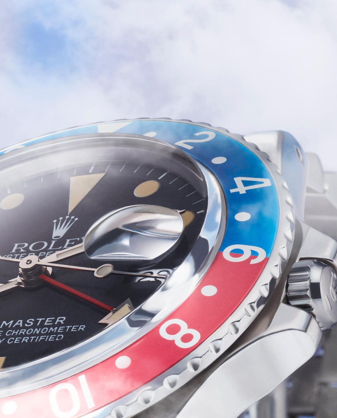 Close-up of a stainless steel Rolex GMT-Master II watch with a black dial and a blue and red bezel against a bright, cloudy sky.