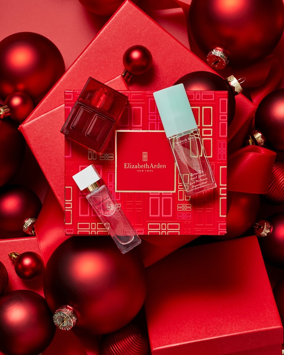 Product shot of three Elizabeth Arden fragrance bottles on a patterned red gift box surrounded by red Christmas ornaments and gift boxes.