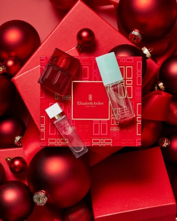 Product shot of three Elizabeth Arden fragrance bottles on a patterned red gift box surrounded by red Christmas ornaments and gift boxes.