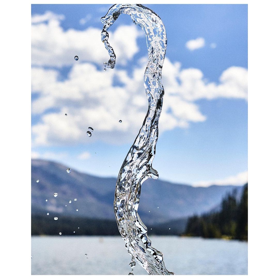 Dynamic shot of clear water stream against a backdrop of lake, mountains, clouds, and blue sky on a sunny day, creating a refreshing and scenic image.