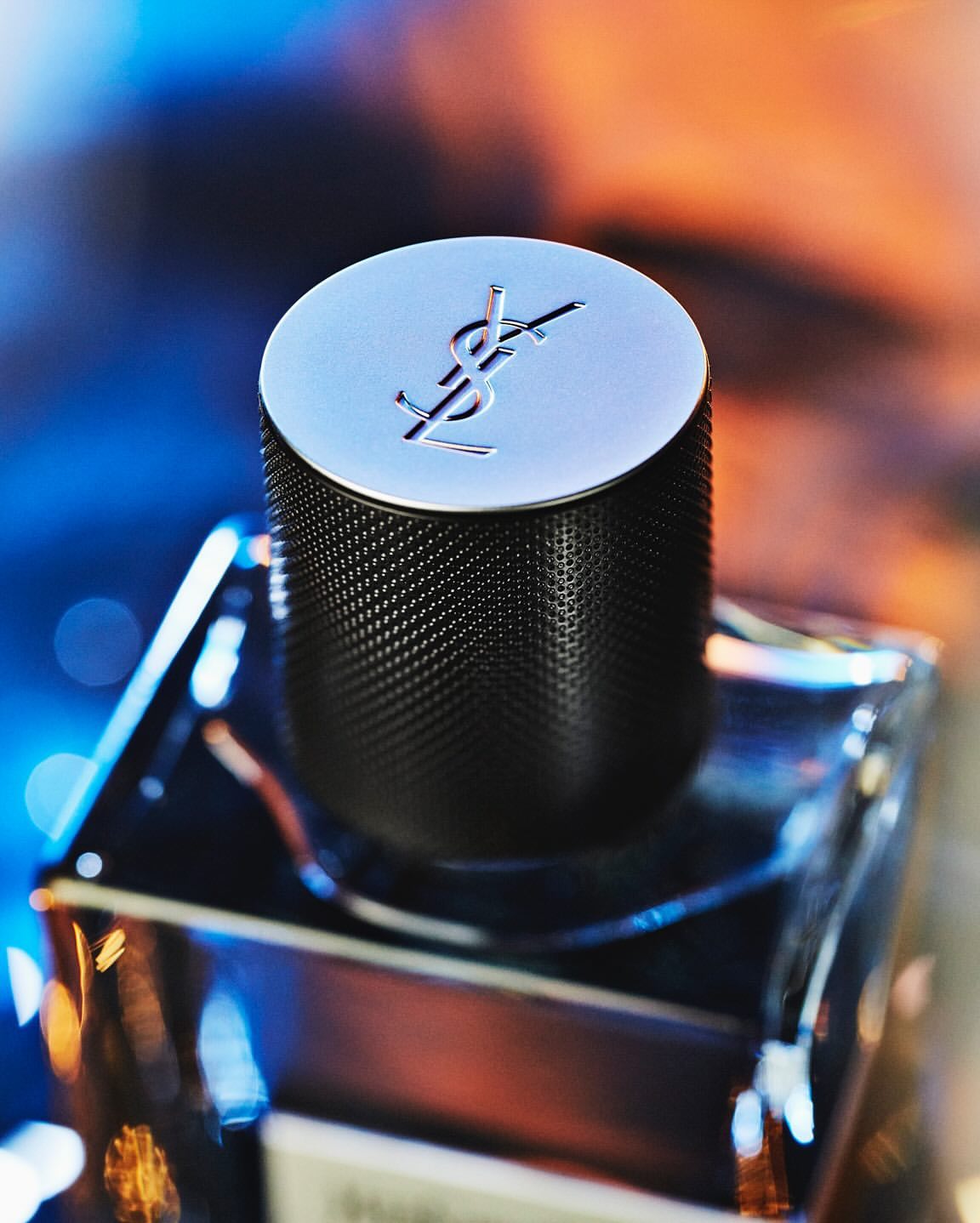 Close-up of a YSL men's fragrance bottle, highlighting the silver cap with the YSL logo, against a bokeh background of blue and orange.