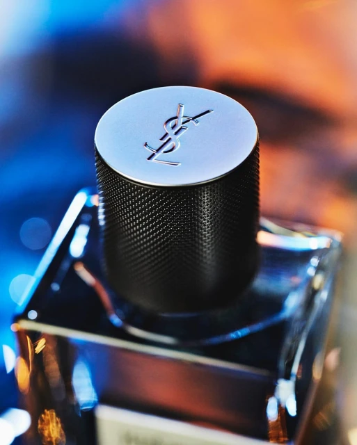 Close-up of a YSL men's fragrance bottle, highlighting the silver cap with the YSL logo, against a bokeh background of blue and orange.