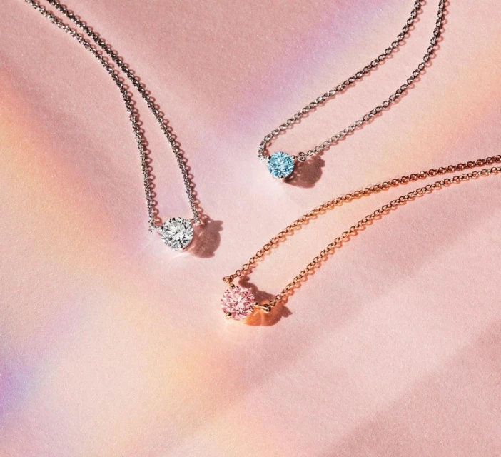 Three delicate necklaces with blue, white, and pink gemstones are displayed on a soft pink surface with rainbow color gradients. 