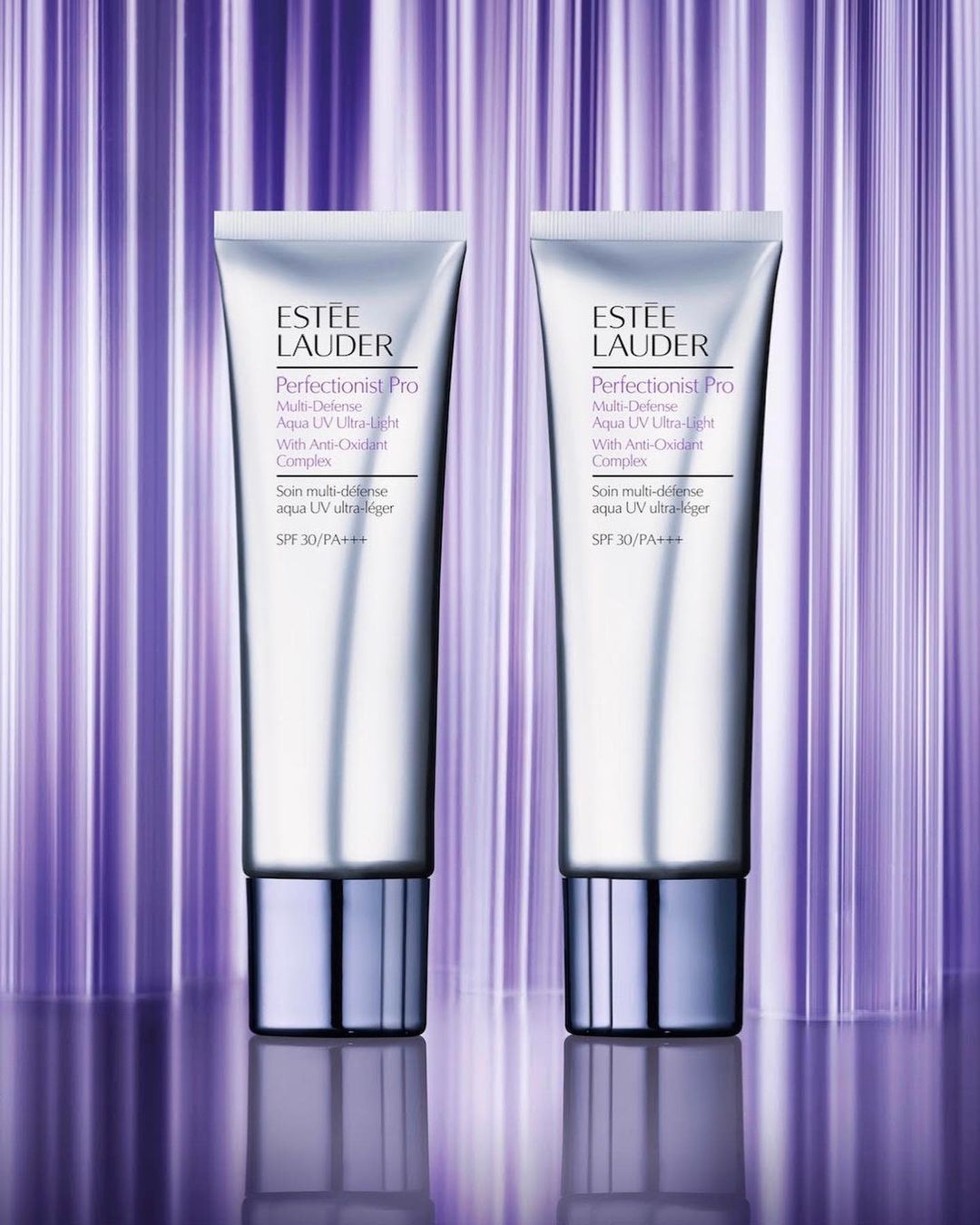 Two silver Estée Lauder Perfectionist Pro Aqua UV Ultra-Light sunscreens with SPF 30 against a striped purple background. Cosmetic product shot.