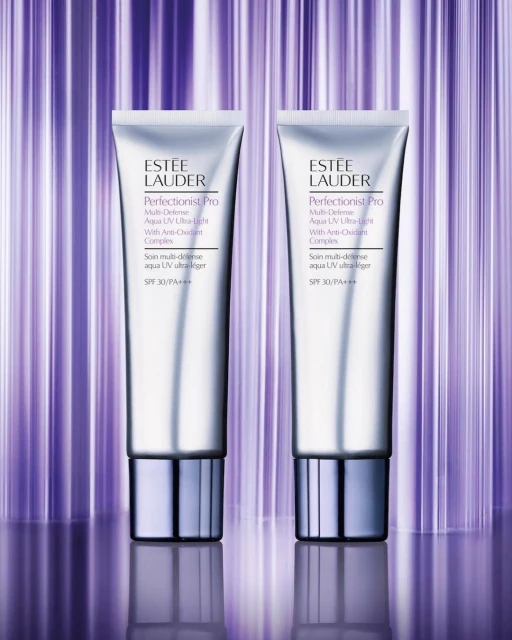 Two silver Estée Lauder Perfectionist Pro Aqua UV Ultra-Light sunscreens with SPF 30 against a striped purple background. Cosmetic product shot.