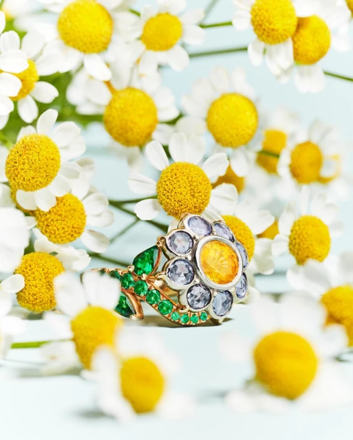Close-up shot of a floral-designed ring featuring yellow and blue stones, nestled among white and yellow chamomile flowers on a light blue surface.