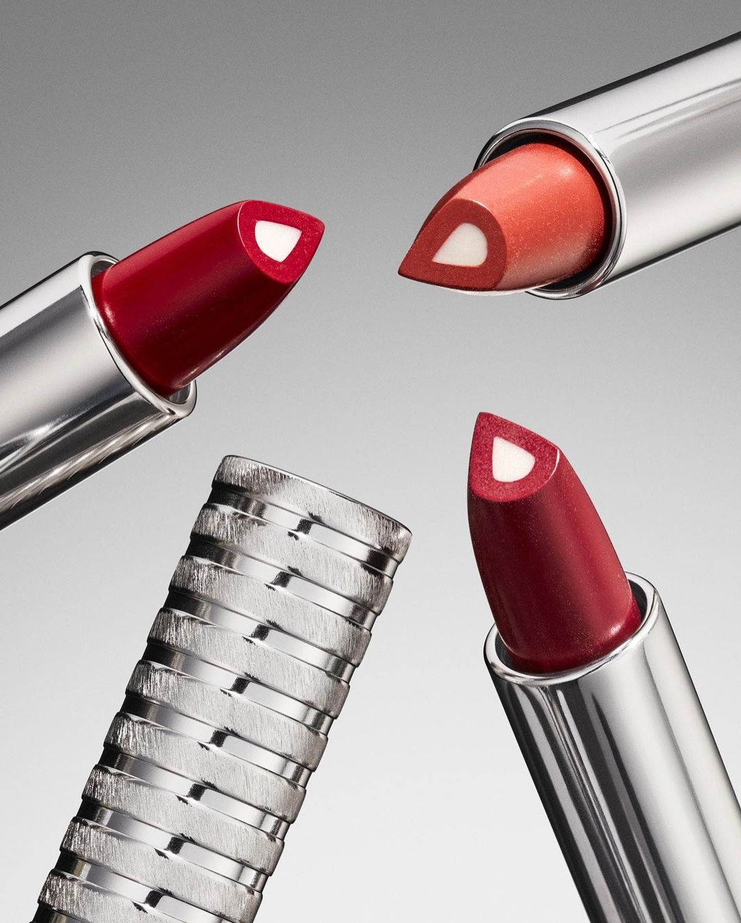 Close-up of three lipsticks with silver tubes and red and coral colors, arranged in a circle against a graduated gray background.