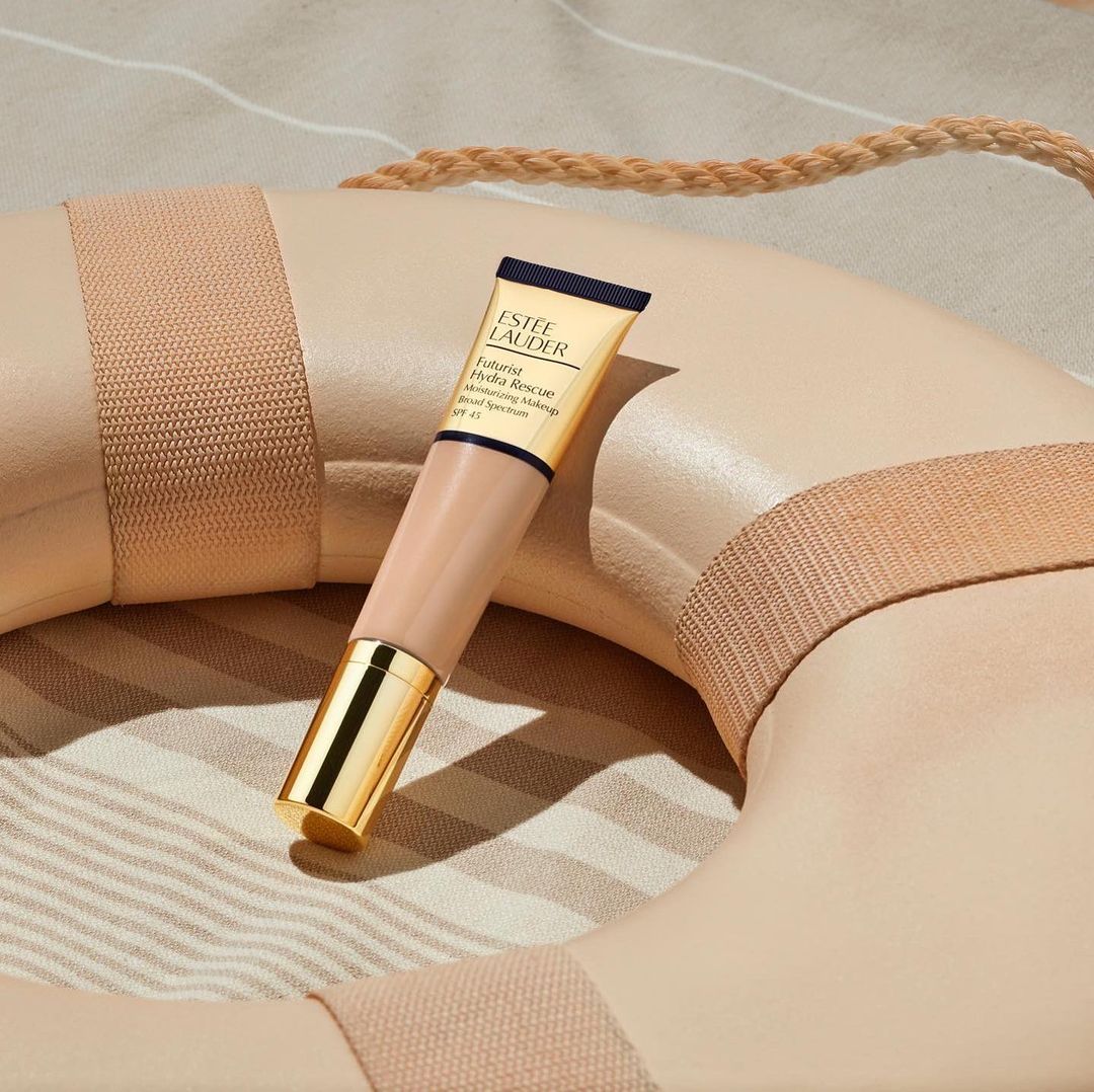 Estee Lauder Futurist Hydra Rescue moisturizing makeup SPF 45 tube rests on a beige life preserver against a striped fabric backdrop.