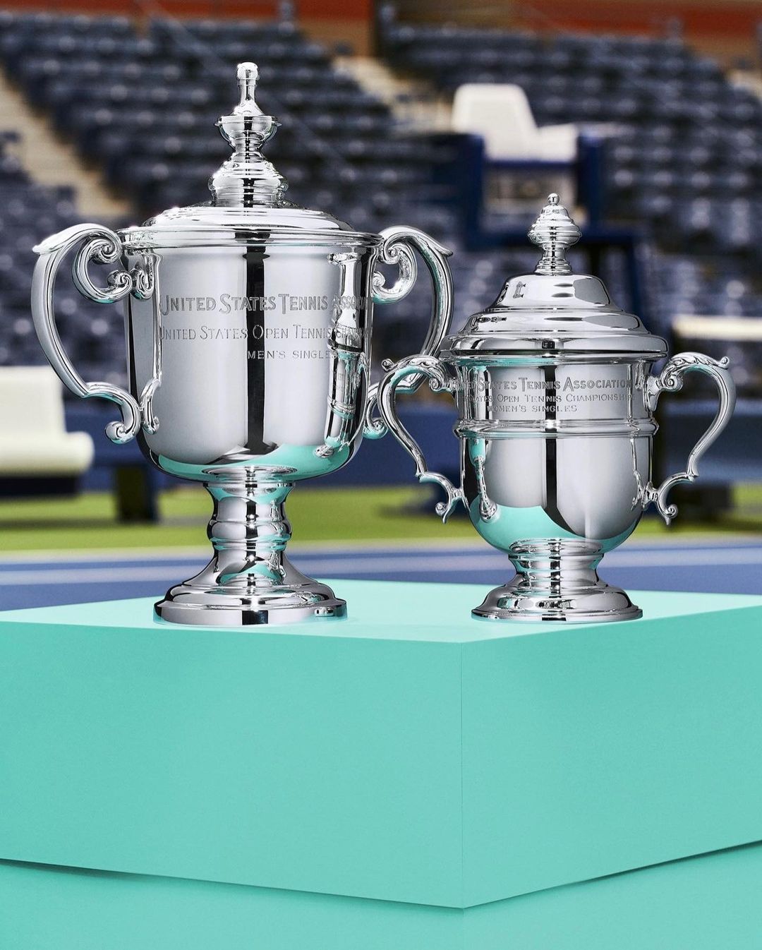 Two United States Tennis Association trophies with lids sit on a turquoise platform against blurred stadium seating backdrop.