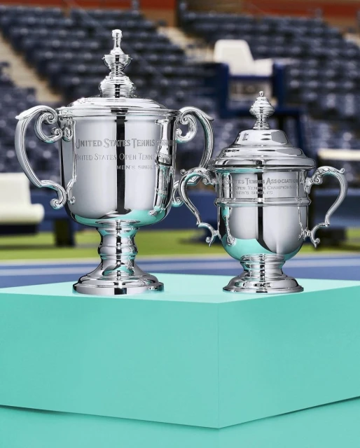 Two United States Tennis Association trophies with lids sit on a turquoise platform against blurred stadium seating backdrop. 