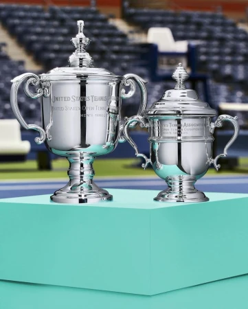 Two United States Tennis Association trophies with lids sit on a turquoise platform against blurred stadium seating backdrop. 