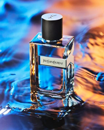 Close-up studio shot of a clear glass Yves Saint Laurent cologne bottle with a black cap, displayed on a reflective surface with blue and orange lighting.