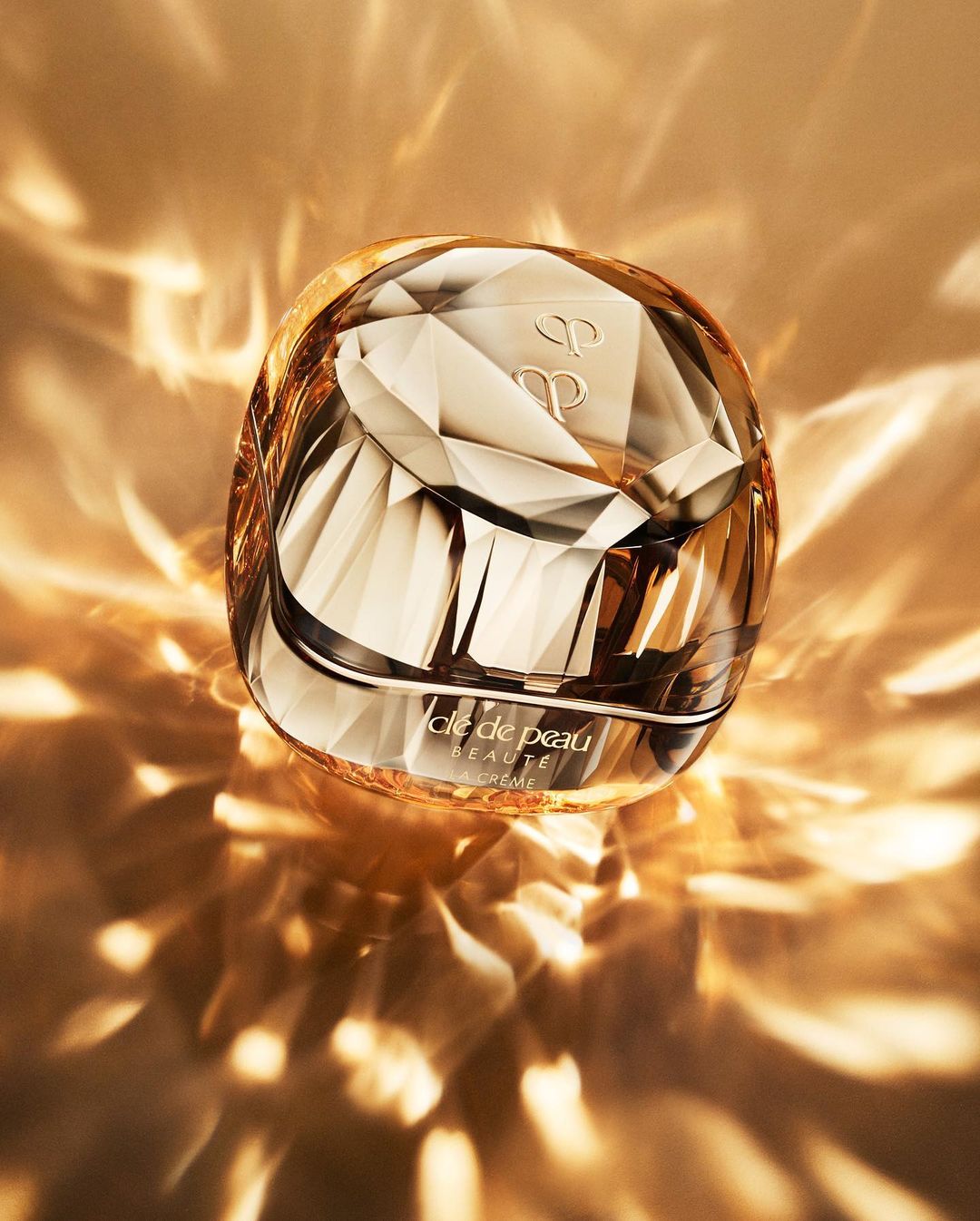 A jar of Clé de Peau Beauté cream sits in a faceted container, reflecting the gold and amber light around it with its geometric design.