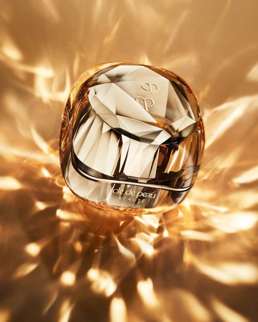 A jar of Clé de Peau Beauté cream sits in a faceted container, reflecting the gold and amber light around it with its geometric design.