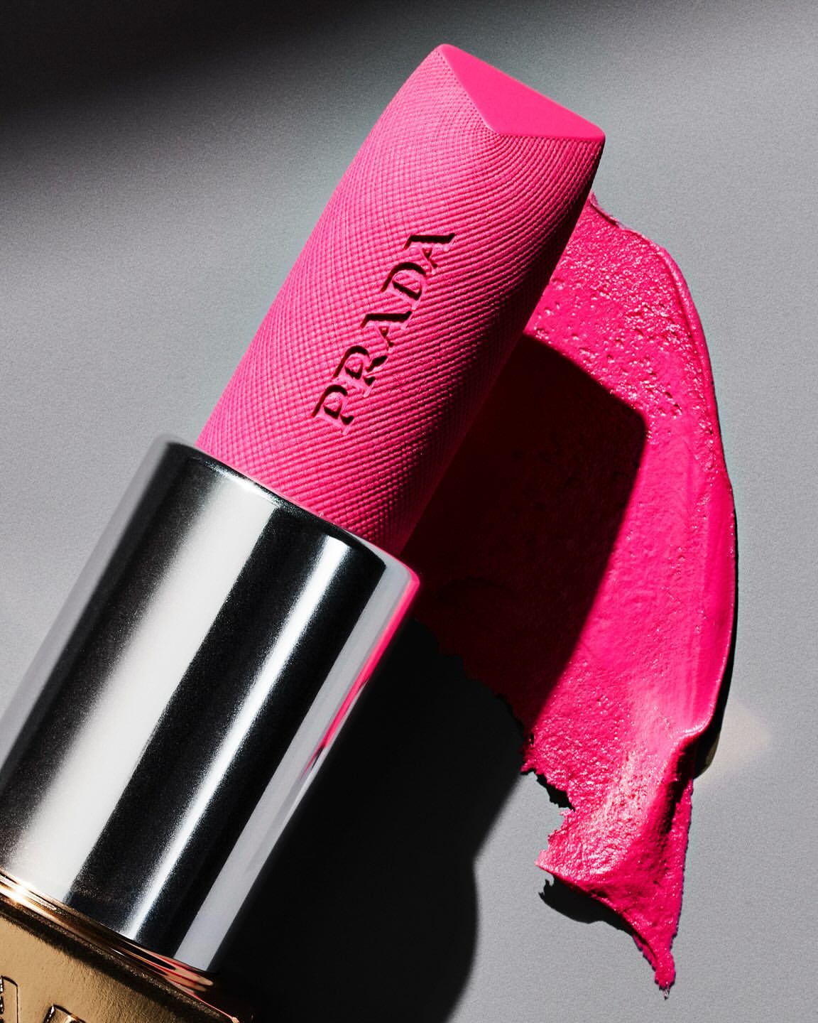Macro shot of vibrant pink Prada lipstick with a silver tube against a gray background, highlighting the texture and color.
