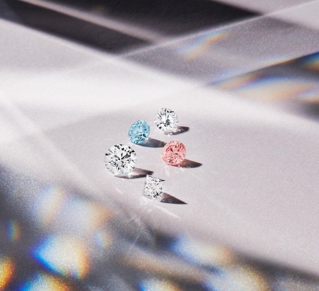 A collection of variously colored and cut diamonds sparkles on a textured white surface with shifting rainbow-colored reflections.