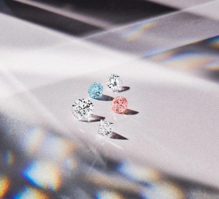 A collection of variously colored and cut diamonds sparkles on a textured white surface with shifting rainbow-colored reflections.