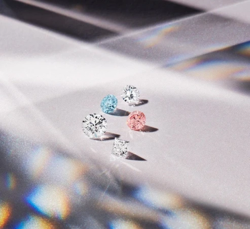 A collection of variously colored and cut diamonds sparkles on a textured white surface with shifting rainbow-colored reflections.
