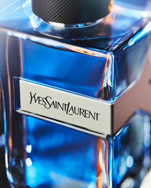 Close-up studio shot of a blue glass Yves Saint Laurent fragrance bottle with a silver label and a black cap against a blurred background.