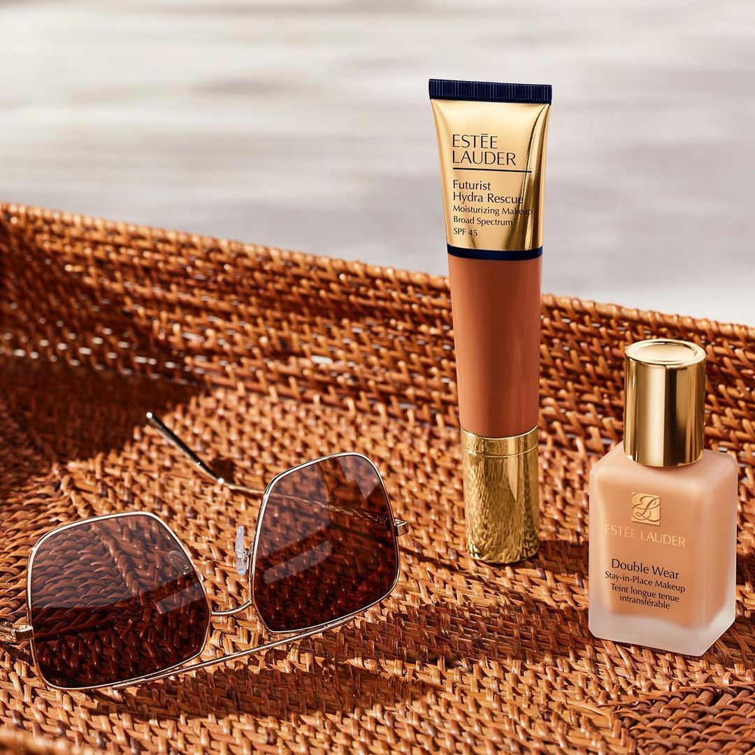 Close-up studio shot of Estee Lauder Futurist Hydra Rescue and Double Wear foundation products with sunglasses on a woven surface.