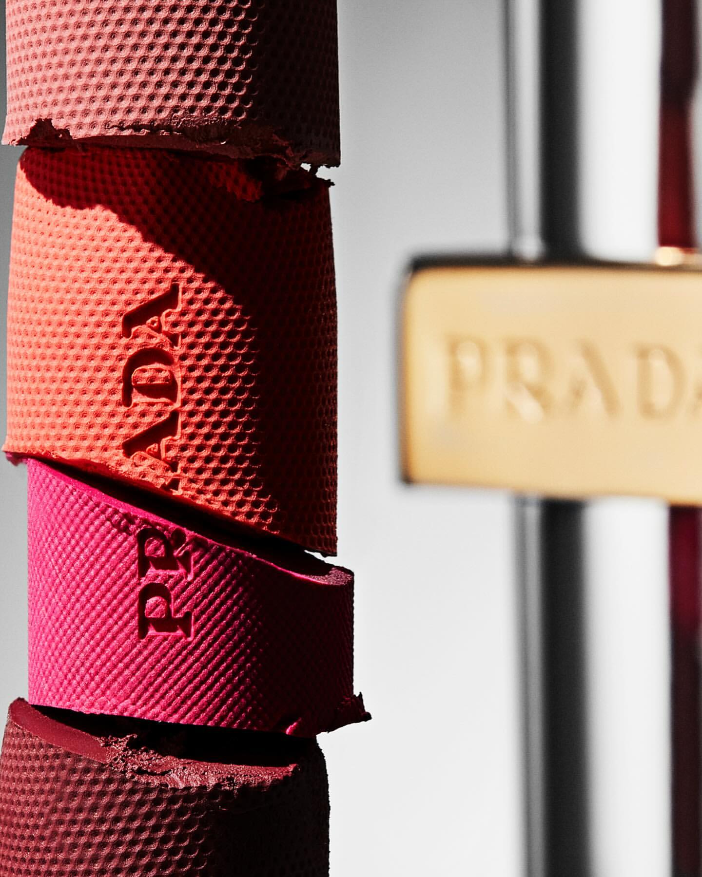 Macro still life shot of stacked Prada Monolith blush and highlighter cases in red, pink, and gold tones with embossed logo details.