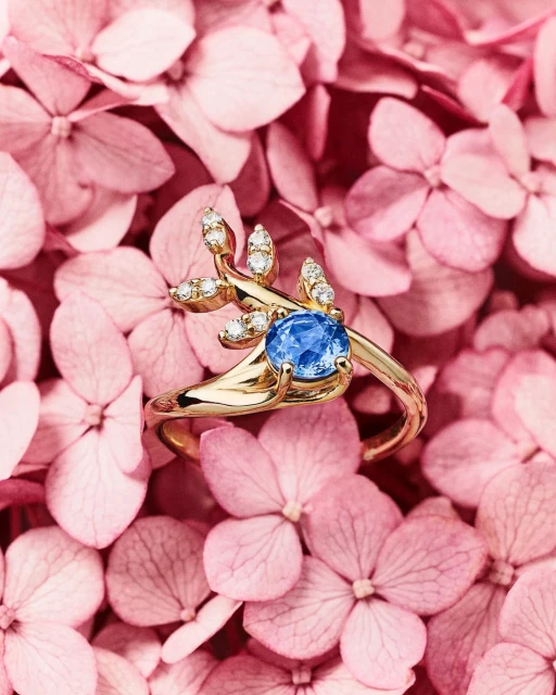 Close-up of a gold ring featuring a blue gemstone and diamond accents displayed on a bed of light pink hydrangea petals.