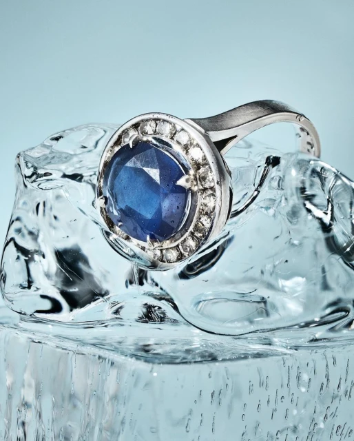 Close-up studio shot of a silver ring with a large blue stone, surrounded by small diamonds, resting on a clear ice cube against light-blue.