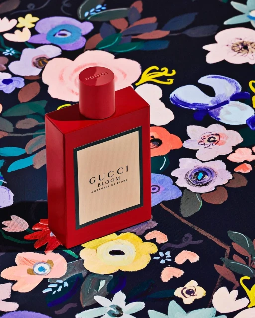 Red Gucci Bloom Ambrosia di Fiori perfume bottle stands out against a painterly floral backdrop featuring diverse colors and blooms.