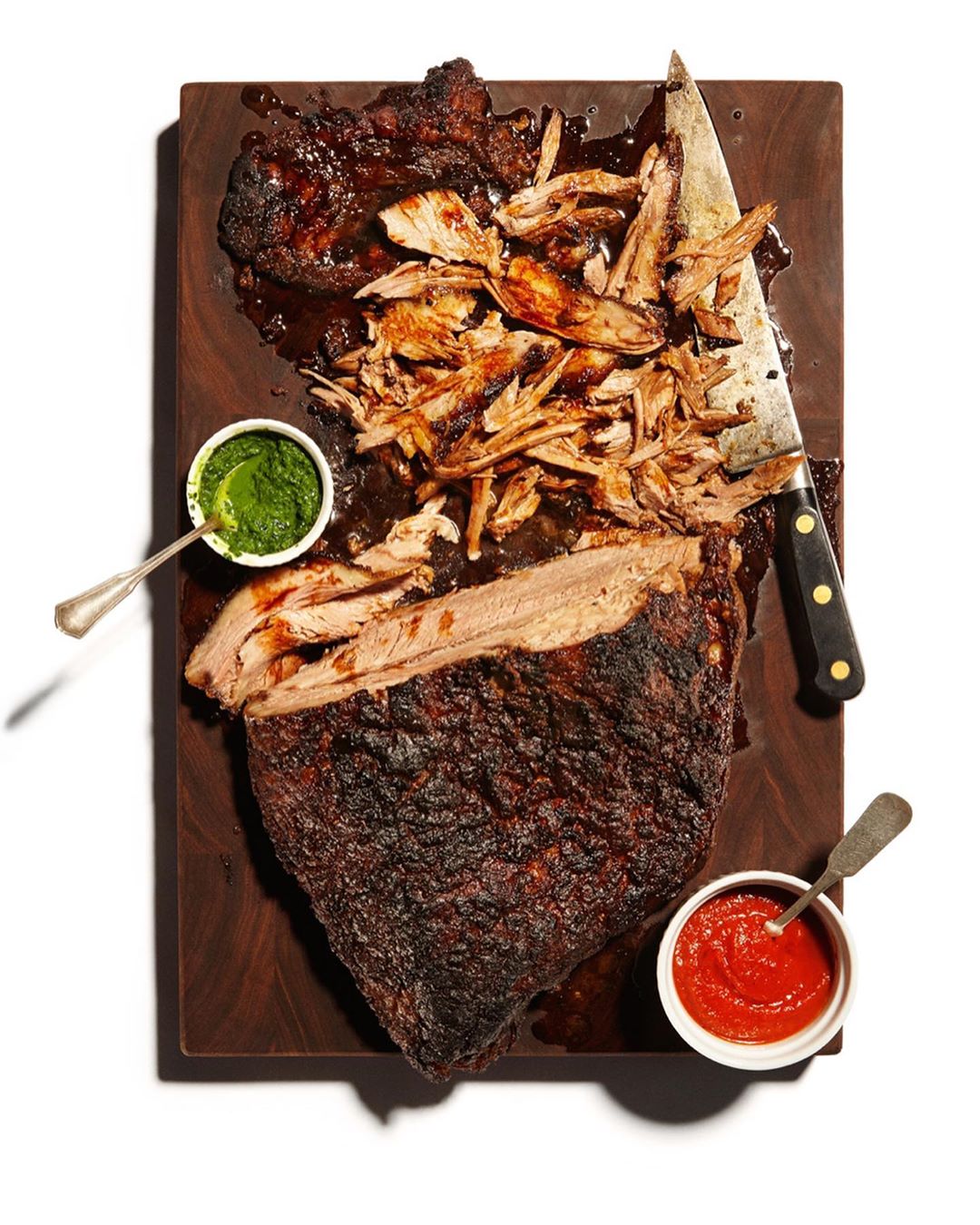 Overhead shot of sliced and whole cooked brisket on a wooden cutting board with small bowls of red and green sauce.