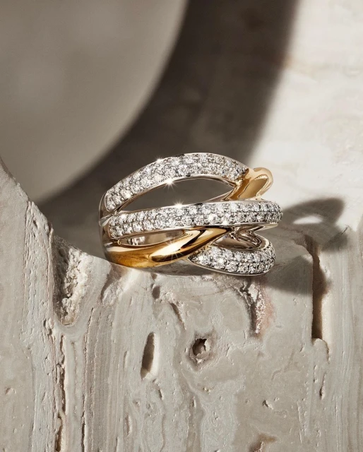 Close-up of a modern statement ring featuring interconnected bands of gold and diamond pavé, resting on a textured surface.