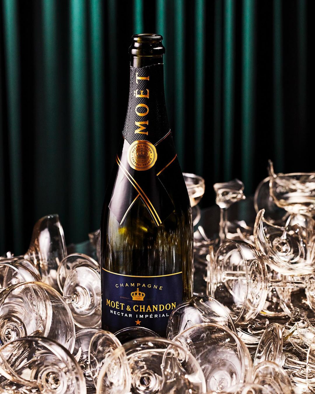 Moet & Chandon champagne bottle in a pile of clear champagne glasses in front of a dark green curtain backdrop.