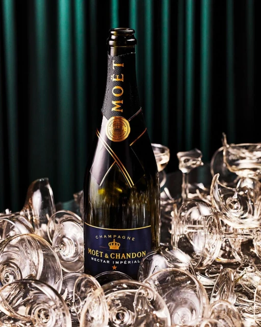 Moet & Chandon champagne bottle in a pile of clear champagne glasses in front of a dark green curtain backdrop.