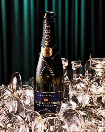 Moet & Chandon champagne bottle in a pile of clear champagne glasses in front of a dark green curtain backdrop.