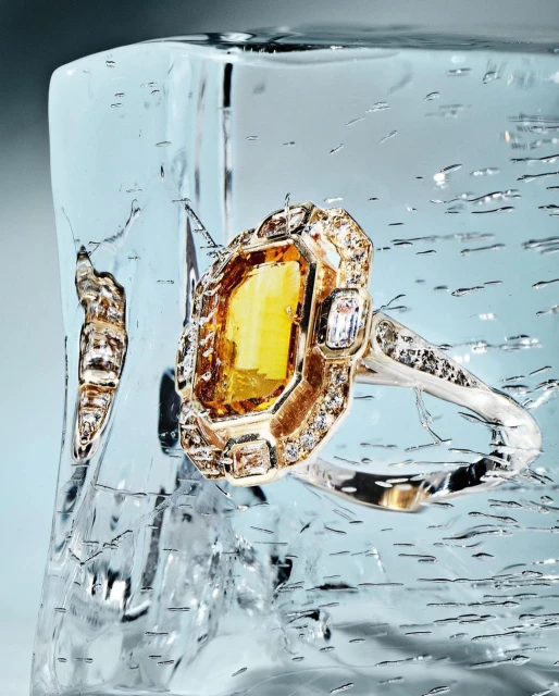 A golden ring with a yellow gemstone is embedded in clear ice, showcased against a light blue backdrop. The ice has cracks and water droplets.