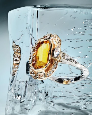 A golden ring with a yellow gemstone is embedded in clear ice, showcased against a light blue backdrop. The ice has cracks and water droplets.