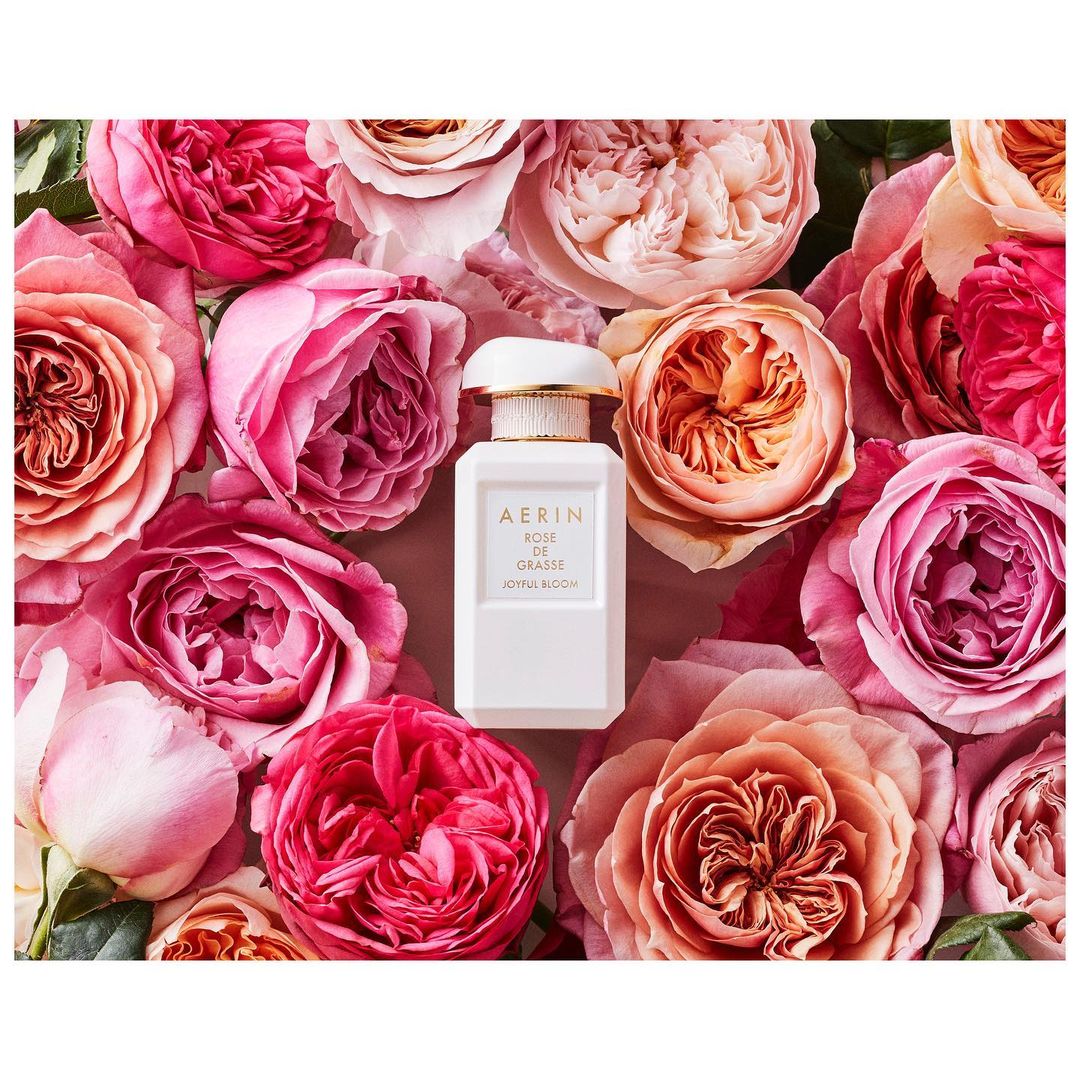 White perfume bottle with gold cap by Aerin, 'Rose de Grasse' scent, surrounded by pink, peach, and cream rose blossoms.