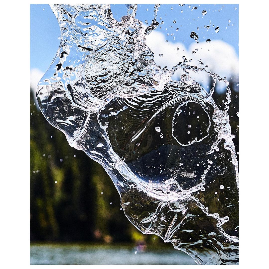 Clear splash of water in mid-air against an outdoor backdrop of green trees and a partly cloudy blue sky on a sunny day.