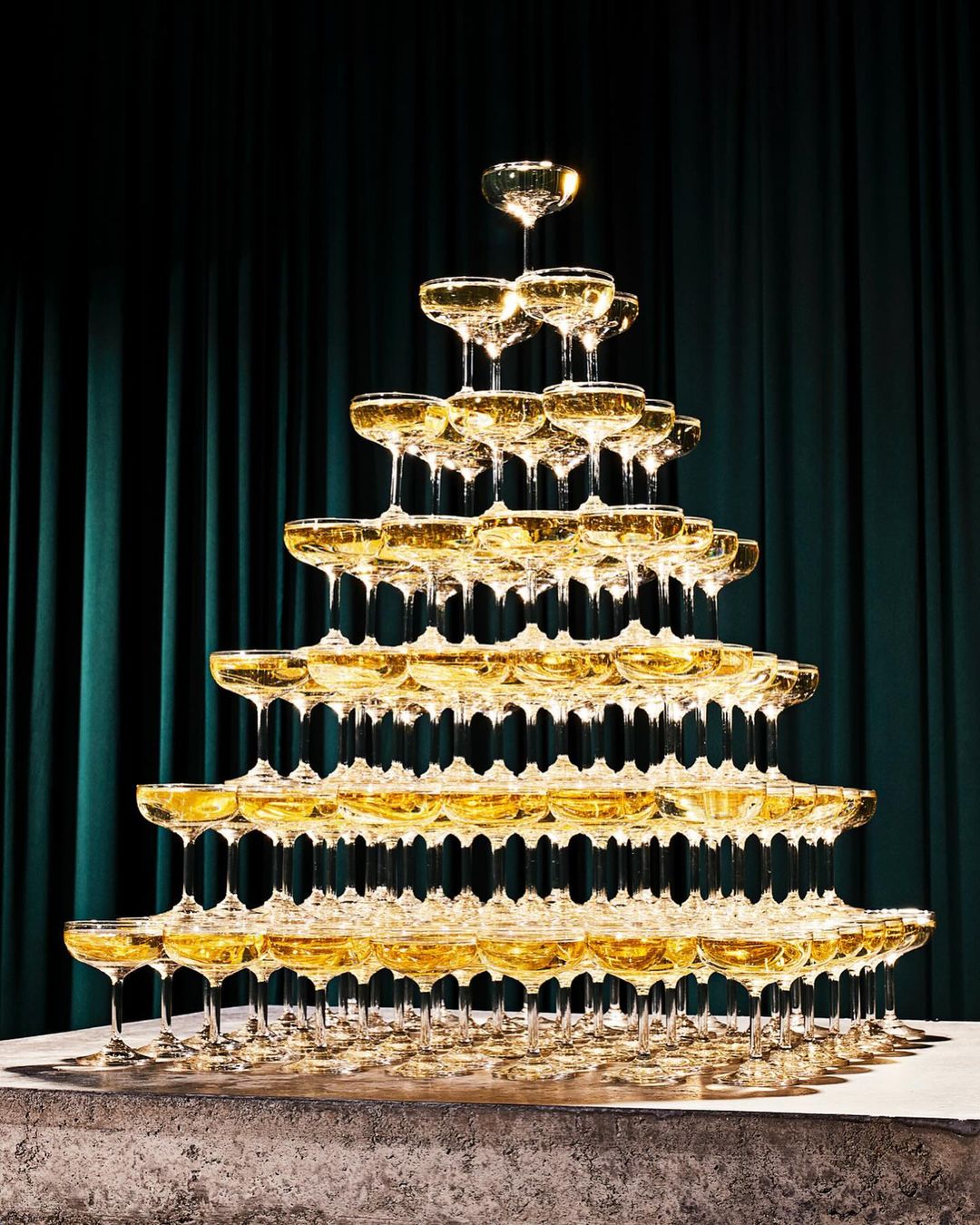 Champagne tower with many glasses filled with sparkling wine stacked high in a pyramid shape, against a draped dark green background.