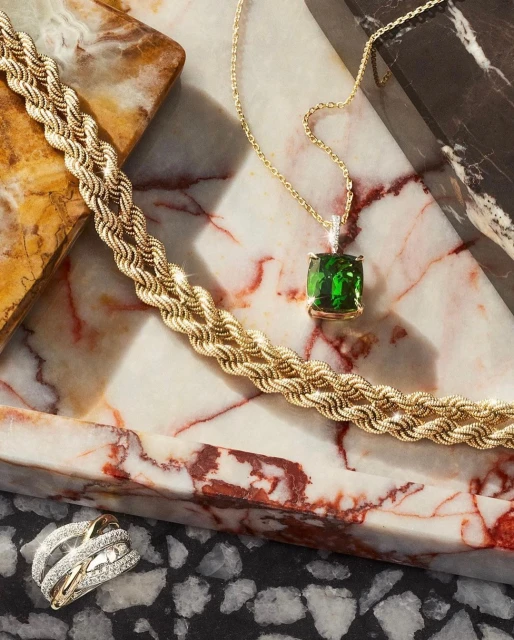 Elegant gold jewelry set on a marble tray. Features a necklace with a green gemstone, a twisted rope chain bracelet, and a modern ring.
