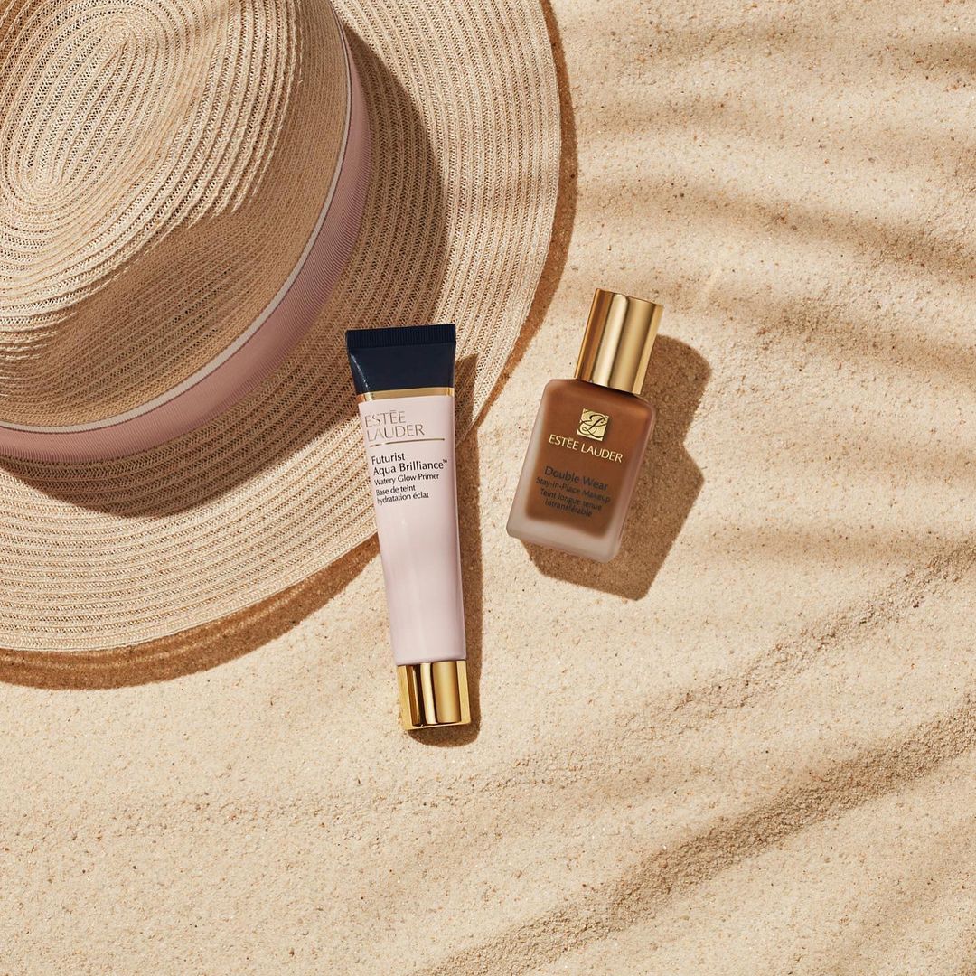 Two Estee Lauder beauty products, a primer and foundation, are staged next to a straw hat on a sandy surface with shadow patterns.
