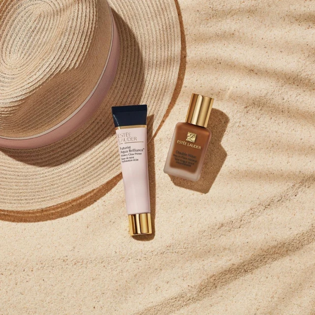 Two Estee Lauder beauty products, a primer and foundation, are staged next to a straw hat on a sandy surface with shadow patterns.
