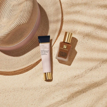 Two Estee Lauder beauty products, a primer and foundation, are staged next to a straw hat on a sandy surface with shadow patterns.