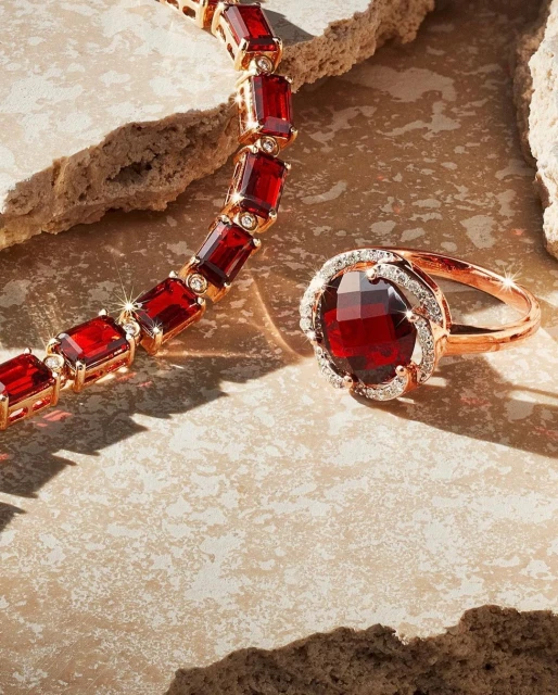 Elegant red gemstone ring with diamonds and gold necklace, set against beige textured stone, perfect for jewelry and fashion advertising.