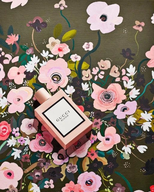 Gucci Bloom perfume bottle on a floral painted surface featuring pink, white, and green flowers against an olive green background.