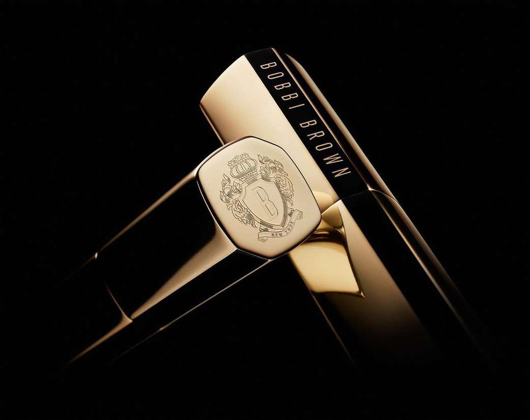 Close-up product shot featuring Bobbi Brown lipstick tubes in gold and black against a dark background, highlighting the brand's logo.