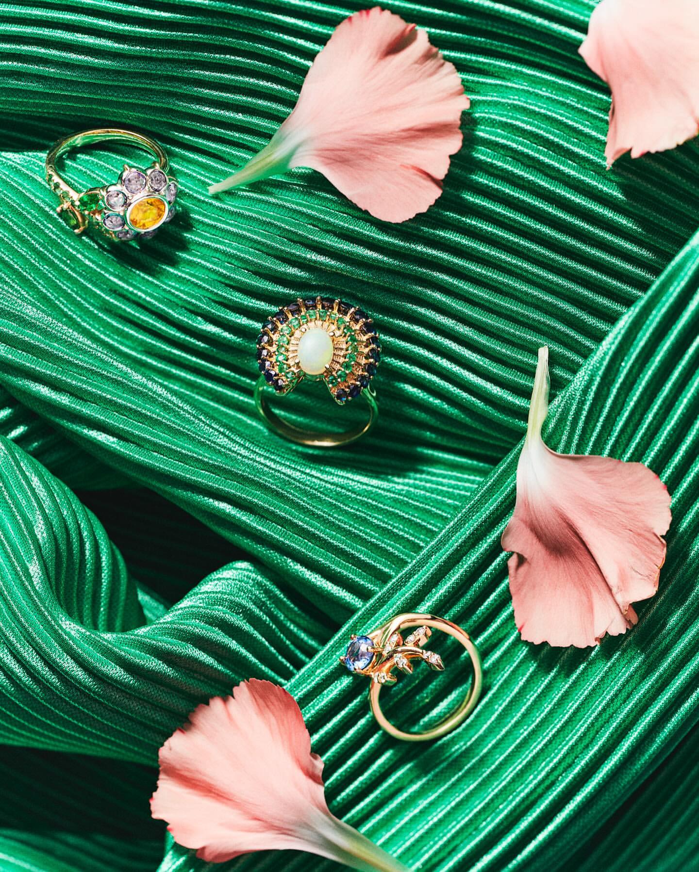 Close-up of three gold rings with gemstones, arranged with pink petals on a textured green fabric, creating an elegant jewelry still life.